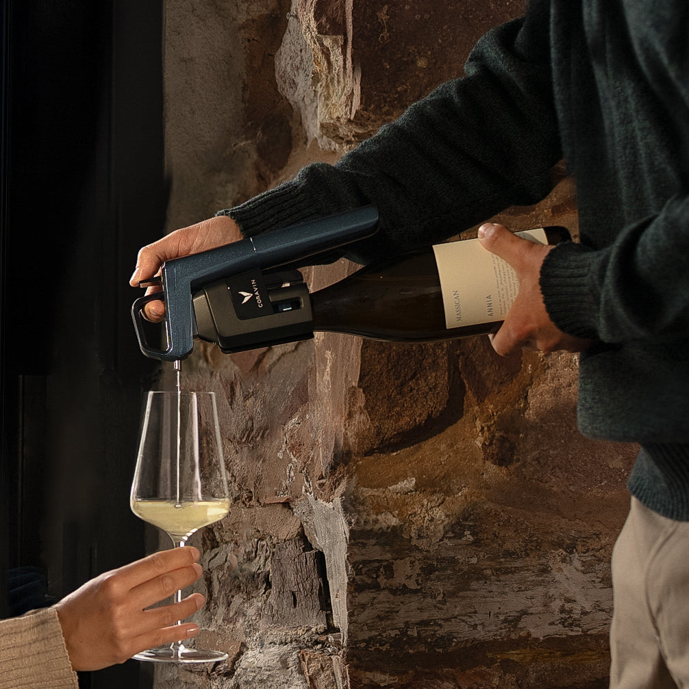 A person pours white wine from a bottle using Corvain Ocean Blue into a glass held by another person, set against a rustic stone wall background.