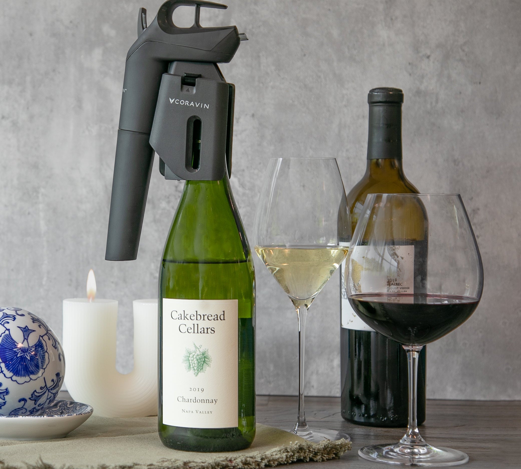 Wine bottles, glasses, and a coravin timeless wine preservation system on a table with a gray background